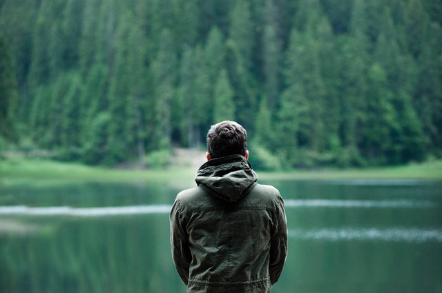 Muž v zelenej bunde stojí pred jazerom vedľa lesa